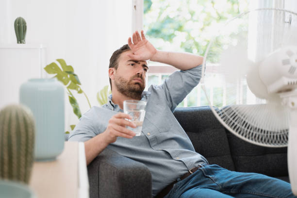 A man wearing a gray shirt sits on a couch while holding a glass of water in his left hand and placing the back of his right hand against his forehead. A portable white fan is placed in front of him in an attempt to cool him down.