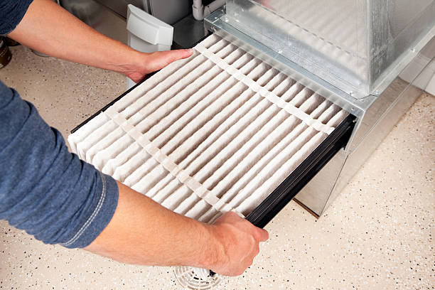 A person with rolled-up sleeves grasps the sides of a furnace filter as they pull it out of the blower compartment.